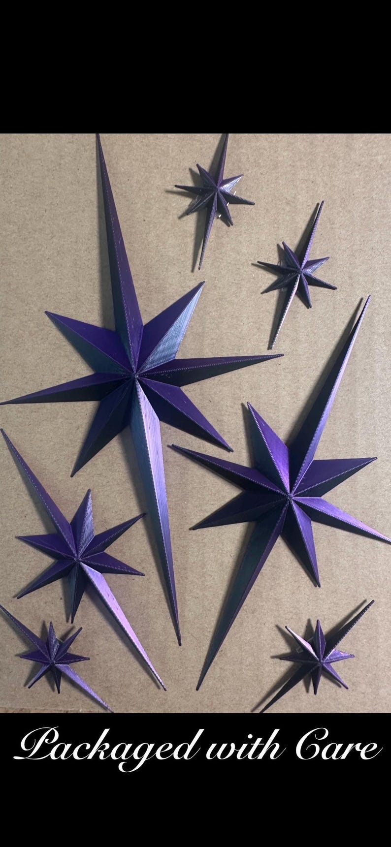 May include: An assortment of purple paper star ornaments in various sizes. The stars have sharp points and are arranged on a neutral background. The text "Packaged with Care" is at the bottom.