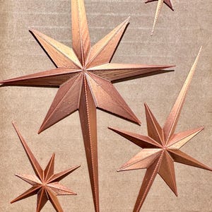 May include: Copper-colored starburst decorations in various sizes. The stars have pointed tips and a textured surface, creating a dimensional effect. The stars are arranged on a neutral background.