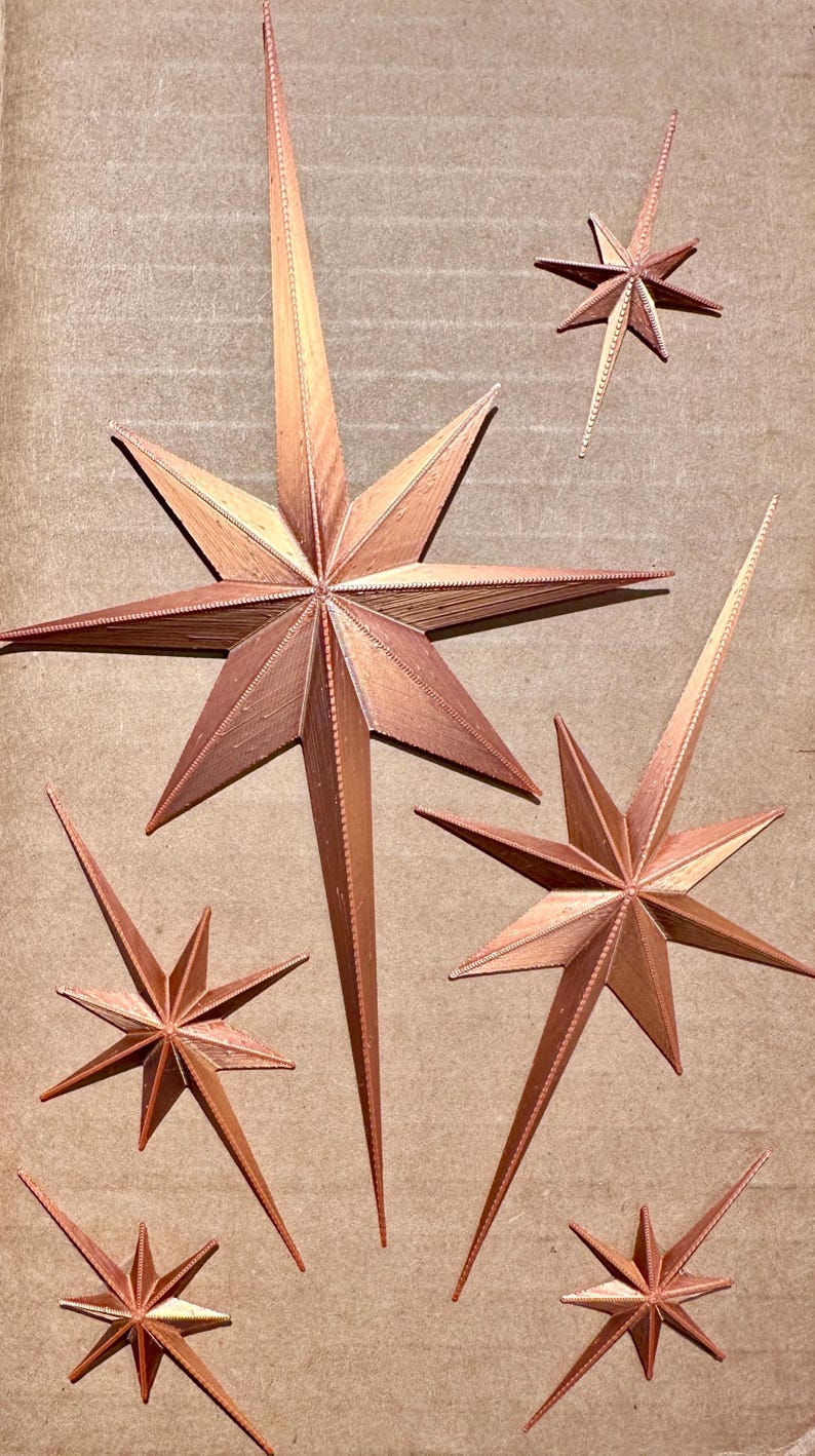 May include: Copper-colored starburst decorations in various sizes. The stars have pointed tips and a textured surface, creating a dimensional effect. The stars are arranged on a neutral background.