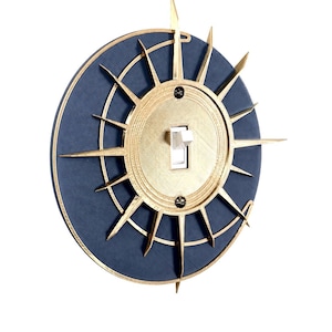May include: A circular light switch cover plate featuring a dark blue textured base and a gold-colored sunburst overlay. The center contains a white toggle switch secured by two screws, with radiating gold spikes extending outward from the center.