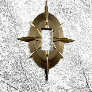 May include: A metallic gold light switch cover plate featuring an oval base with a central rectangular cutout and eight radiating pointed spikes extending from the perimeter, creating a starburst or compass-like design with a textured finish.