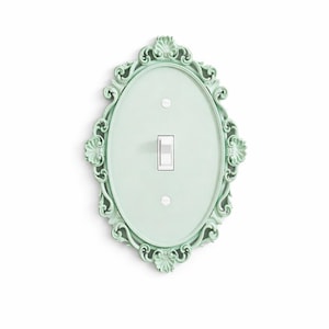 Puede incluir: Una cubierta decorativa para interruptor de luz en un tono verde pálido. La placa ovalada presenta un marco ornamentado con volutas. El interruptor y las cubiertas de los tornillos son blancos. Un acento de decoración del hogar único.
