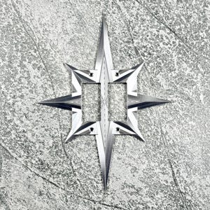 May include: A metallic silver double light switch plate featuring a sharp, eight-pointed star design. The plate has two rectangular cutouts for switches and four small circular screw holes positioned at the top and bottom of the central frame.