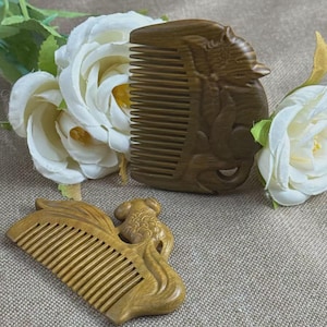 May include: Two wooden combs with carved designs. One comb is a light brown color and features a fish design. The other comb is a darker brown color and features a carved animal design. White roses are in the background.