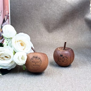 May include: Two small, round, brown wooden apple-shaped ornaments. One apple features a laser-engraved floral wreath with the text "Roxanne & Action". The other apple displays a laser-engraved design of two interlocking rings with a heart.