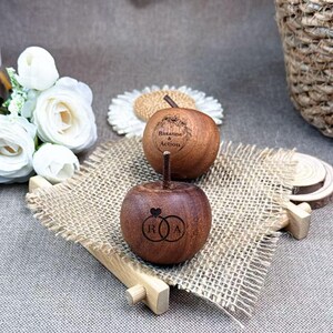 May include: Two wooden apple-shaped boxes with stems, one engraved with "Roxanne & Action", the other with interlocking rings and the letters "R" and "A". They rest on a burlap surface, with white flowers and a woven basket in the background.