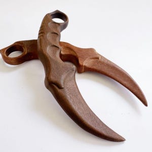 Op de afbeelding: Twee gebogen houten gereedschappen in karambit-stijl met een donkerbruine afwerking, een vingerring aan de bovenkant van het handvat en een voorgevormde grip. De bladen hebben een duidelijke sikkelvorm en tonen de natuurlijke houtnerfstructuur.
