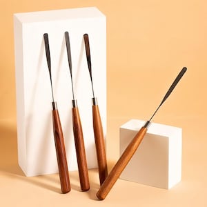 May include: A set of four palette knives featuring long, slender, dark metal blades and smooth, cylindrical wooden handles. Three knives are positioned vertically against a tall white block, while one knife rests at an angle on a smaller white block.
