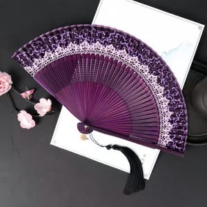 May include: A deep purple folding hand fan featuring a dark purple fabric leaf with a swirling vine pattern and a wide, light pink lace trim along the top edge. The fan has a black tassel attached to the base and dark purple wooden ribs.