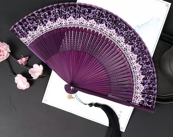 May include: A deep purple folding hand fan featuring a dark purple fabric leaf with a swirling vine pattern and a wide, light pink lace trim along the top edge. The fan has a black tassel attached to the base and dark purple wooden ribs.