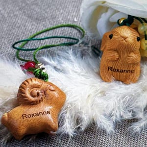 May include: Two wooden pendants, one shaped like a ram and the other like a bunny, both with the name "Roxanne" engraved. The pendants are attached to green cords with red and silver accents. The background is white feathers.