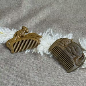 May include: Two wooden combs with carved animal designs. One comb features a goldfish, the other a sleeping fox. Both combs are brown and have fine teeth. The combs are displayed on a textured, neutral-colored surface.