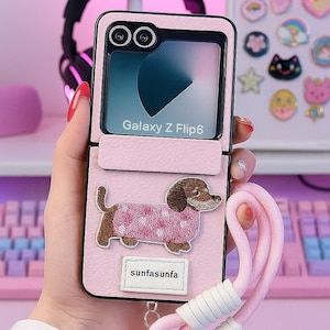 May include: Pink Galaxy Z Flip6 phone case with a dachshund dog design. The case has a matching pink wrist strap with a fluffy pom-pom and the text "sunfasunfa". A pink cat ear headband is in the background.