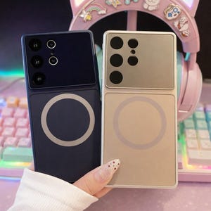 Puede incluir: Dos fundas de teléfono, una azul marino y otra beige, se sostienen frente a unos auriculares rosas con orejas de gato. Las fundas tienen un diseño rectangular con un acento circular. La funda azul marino tiene tres lentes de cámara y la beige tiene seis.