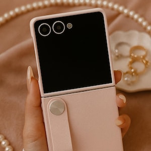 May include: A pink foldable smartphone with a black screen and a camera module. The phone is held in a hand, showcasing its design. The phone has a strap and a silver button. The background includes jewelry and a pearl necklace.