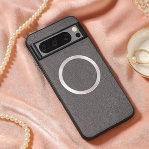 Può includere: Una custodia per telefono grigio scuro con un anello argentato e un modulo fotocamera nero. La custodia è su una superficie di raso rosa, con una collana di perle e una piccola ciotola con un anello sullo sfondo.