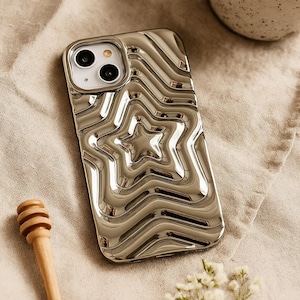 May include: A metallic silver phone case featuring a raised, concentric star pattern with a liquid-like, reflective surface. The design includes a cutout for the camera lens and flash, showcasing a smooth, undulating texture across the entire back.