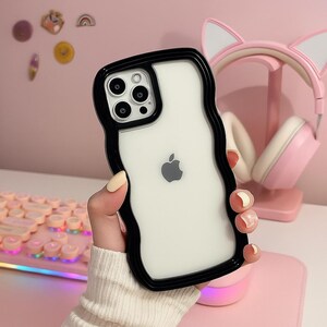 Puede incluir: Una funda transparente para iPhone con un borde negro ondulado, que muestra el logotipo de Apple. La funda se sostiene frente a un fondo rosa, con unos auriculares y un teclado rosas visibles. La funda está diseñada para proteger el teléfono.