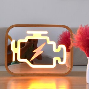 Puede incluir: Un espejo rectangular con marco de madera redondeado que presenta un símbolo luminoso de estilo neón de una luz de control del motor en el centro. La luz amarilla brillante muestra el contorno clásico del motor con un icono de rayo.