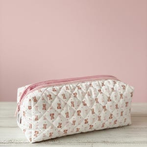 May include: A rectangular, quilted cosmetic bag with a repeating pattern of brown teddy bears and pink bows on a white background. The bag has a pink zipper and a pink and white gingham lining. The bag is on a white wooden surface.