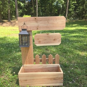 May include: A wooden outdoor sign with a planter box base. The sign has two horizontal planks for text, a small picket fence, and a hanging lantern. The structure is made of light-colored wood and sits on grass.
