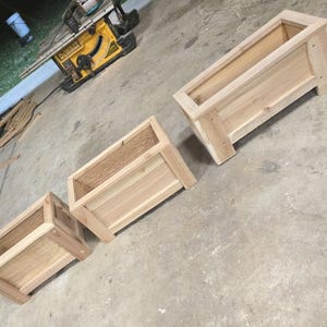 Könnte beinhalten: Drei unfertige rechteckige Holzpflanzkästen in verschiedenen Größen. Die Pflanzkästen sind aus hellfarbenem Holz gefertigt und haben ein einfaches Design mit offener Oberseite. Sie stehen auf einer Betonoberfläche, was auf ein DIY- oder Holzbearbeitungsprojekt hindeutet.