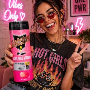 Puede incluir: Un frasco de spray rosa y negro con el texto "Ratchet" y "Witches, Hoes, & Assholes". El frasco lo sostiene una persona que lleva una camiseta negra con un gráfico y las palabras "Hot Girls Don't Bug".