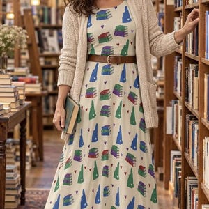 May include: A cream-colored maxi dress featuring a repeating pattern of stacked books, green and blue state silhouettes, and small red hearts. The dress is styled with a beige open-front knit cardigan and a brown leather belt with a metal buckle.