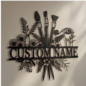 Puede incluir: Un letrero de pared de metal negro con el texto CUSTOM NAME centrado horizontalmente. El diseño incluye siluetas de tres pinceles que apuntan hacia arriba y hacia abajo, rodeados por varios elementos botánicos florales y de hojas.