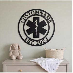May include: A circular black metal wall sign featuring the text CUSTOMNAME at the top and EST. 2023 at the bottom. In the centre, a black Star of Life symbol is displayed, which includes a staff with a serpent wrapped around it.