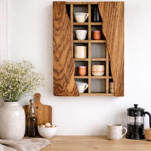 Peut inclure: Une étagère murale en bois avec une grille de compartiments, contenant diverses tasses et petits objets décoratifs. L'étagère est en bois brun clair et est fixée sur un mur blanc. Un comptoir de cuisine avec un vase de fleurs, une planche à découper et une cafetière est au premier plan.