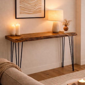 Peut inclure: Une console rectangulaire dotée d'un plateau en bois brut à bord naturel avec une finition brun riche, reposant sur quatre pieds épingle fins en métal noir. La table est placée contre un mur, supportant deux bougies blanches et une lampe.