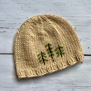 May include: A beige knitted beanie with a green embroidered design of three pine trees.