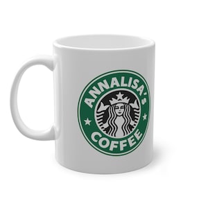 Può includere: Tazza da caffè in ceramica bianca con un logo verde e bianco che recita "Annalisa's Coffee". Il logo è una parodia del logo Starbucks, con il volto di una donna e le parole "Annalisa's Coffee" in un design circolare.