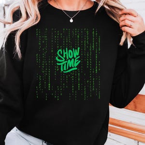 May include: Black sweatshirt with the words "SHOW TIME" in green, stylized text. The design includes a matrix-style background of green numbers. The sweatshirt is a casual, comfortable garment.