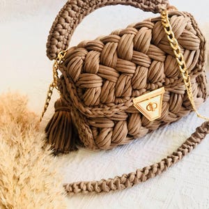 Pode incluir: Uma mala de mão castanha tecida com uma alça de corrente dourada e uma pega. A mala apresenta um fecho dourado e uma borla. A mala é feita de um material macio e texturizado, adequado para uso diário.