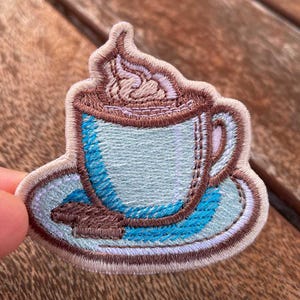 May include: Embroidered patch depicting a coffee cup with whipped cream. The cup and saucer are light blue with brown detailing. The patch has a stitched border and is designed to be ironed on.