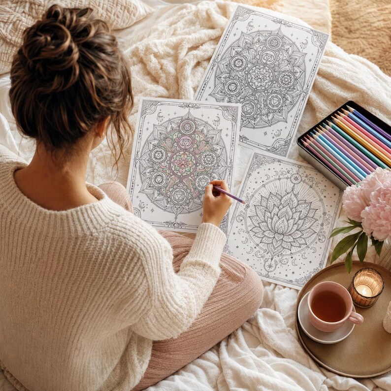 May include: Three white paper sheets featuring intricate black line art mandala and lotus flower designs, a metal tin containing multicolored pencils, and a pink mug on a tray. One sheet is partially colored with soft pastel shades.