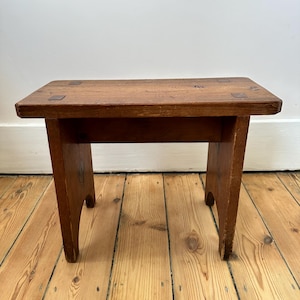 Può includere: Un piccolo sgabello rettangolare in legno con finitura naturale. Lo sgabello ha quattro gambe e un piano piatto. Le gambe sono leggermente curve. Lo sgabello è in legno e ha un aspetto rustico.