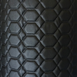 May include: Close-up of a black, textured material with a repeating hexagonal pattern. The pattern is quilted, creating a raised, padded effect. The material appears to be leather or a leather-like fabric, with a slightly pebbled surface.