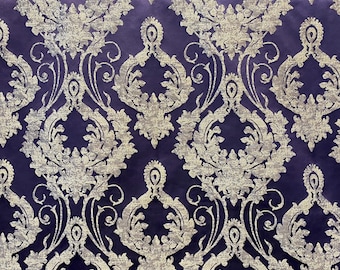 May include: Close-up of a luxurious fabric with a damask pattern. The fabric features a deep purple background with a silver-colored, ornate floral design. The pattern is symmetrical and detailed, creating a rich, elegant texture.