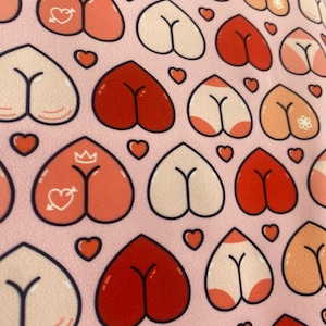 May include: Pink fabric with a repeating pattern of heart-shaped buttocks in various shades of red, peach, and white, outlined in black. Small orange hearts are scattered throughout the design.
