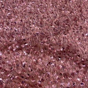 May include: Close-up of a textured, pink fabric with a fluffy, shaggy appearance. The fabric is densely covered with small, round, shiny sequins that catch the light. The overall effect is a soft, shimmering texture, suitable for apparel or decorative projects.