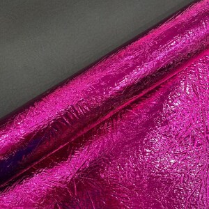 Pu&ograve; includere: Un rotolo di tessuto rosa acceso vibrante caratterizzato da una finitura metallica altamente riflettente con una superficie distintamente stropicciata e testurizzata. Il materiale mostra una lucentezza brillante che cattura la luce.