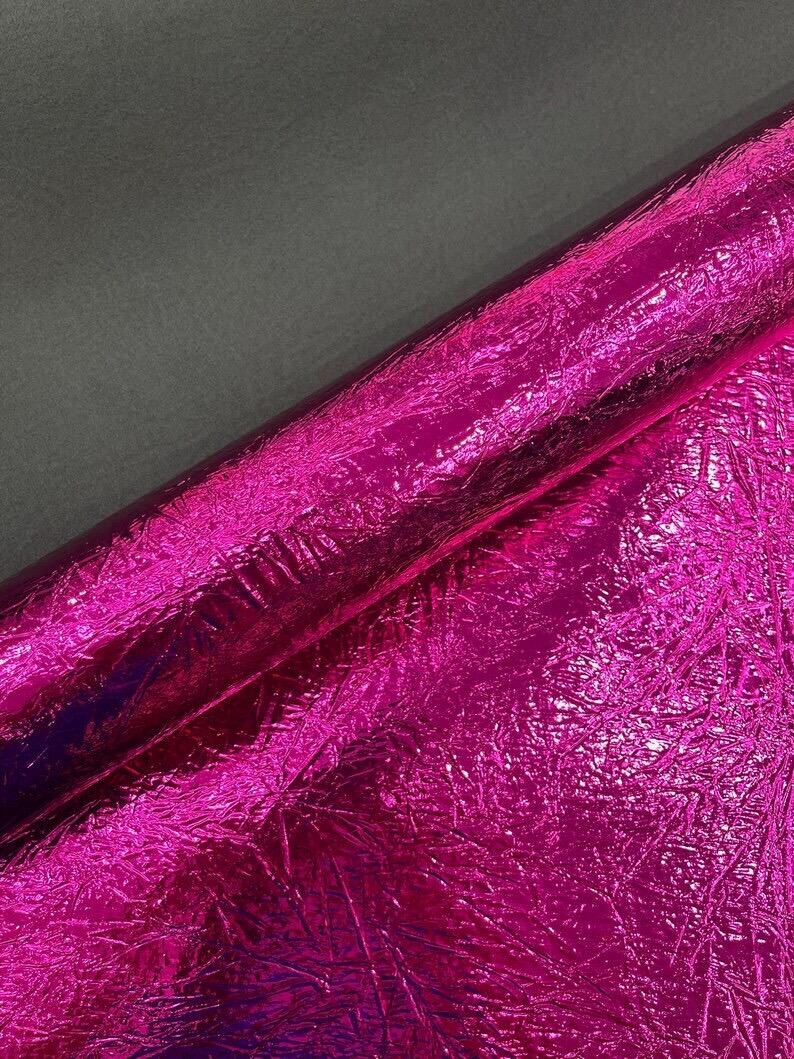 Pu&ograve; includere: Un rotolo di tessuto rosa acceso vibrante caratterizzato da una finitura metallica altamente riflettente con una superficie distintamente stropicciata e testurizzata. Il materiale mostra una lucentezza brillante che cattura la luce.