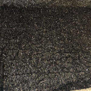 May include: Close-up of a black fabric with a textured surface. The fabric is covered in small, shiny, silver sequins, creating a sparkling effect. The material appears soft and could be used for apparel or decorative purposes.