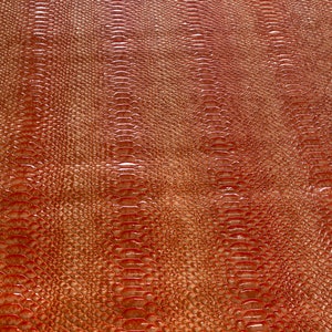 May include: Close-up of a textured, reddish-brown material with a reptile-like scale pattern. The surface has a glossy finish, reflecting light and creating highlights. The design resembles snake or alligator skin, suitable for fashion or craft projects.