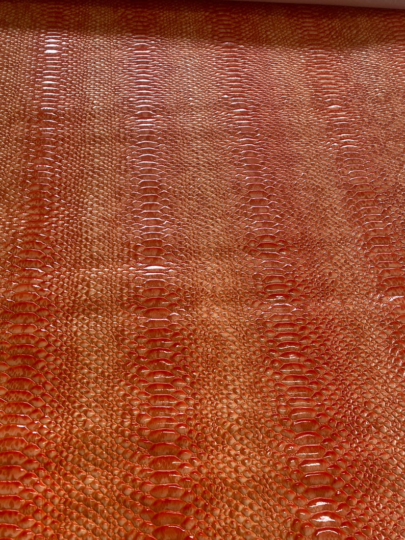 May include: Close-up of a textured, reddish-brown material with a reptile-like scale pattern. The surface has a glossy finish, reflecting light and creating highlights. The design resembles snake or alligator skin, suitable for fashion or craft projects.