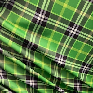 May include: Green and black plaid fabric with a subtle sheen. The fabric is folded and draped, showing the texture and pattern.