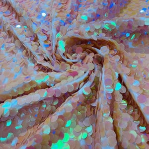 May include: A fabric covered in round, peach-colored sequins that feature an iridescent finish. The sequins are densely packed and overlap, creating a shimmering, reflective surface with subtle flashes of blue and green light across the texture.